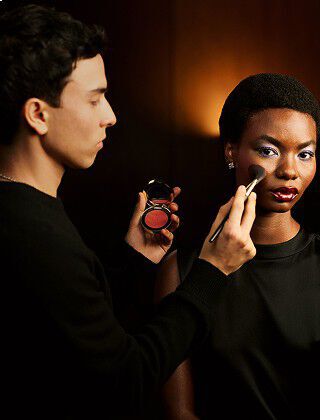 Makeup artist Sam Visser applies blush to a model with a bold eye look and glossy red lips, holding a YSL blush compact.