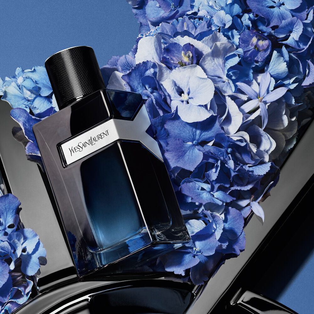 YSL Y Eau de Parfum bottle on a glossy black YSL logo, surrounded by vibrant blue hydrangeas, against a deep blue background.