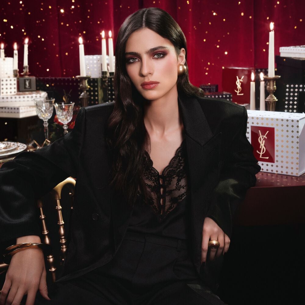 Model wearing a black suit and glam look, seated in a festive candlelit setting with YSL gift boxes.