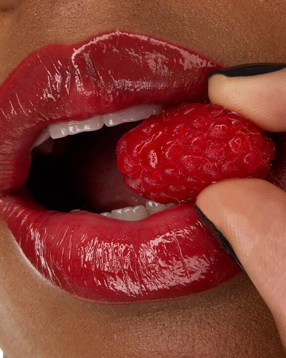 Close-up of model Zahara's lips, adorned with YSL LOVESHINE Lipstick in Berry Crush, shade 219, holding a fresh raspberry.