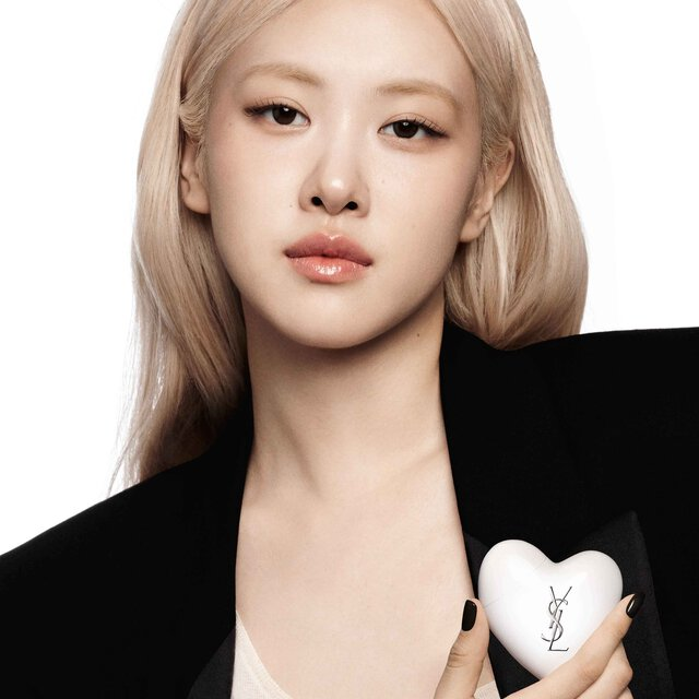 Model in a black leather jacket holding Care Crush Hand & Body serum-in-cream, a white heart-shaped accessory on a chain.